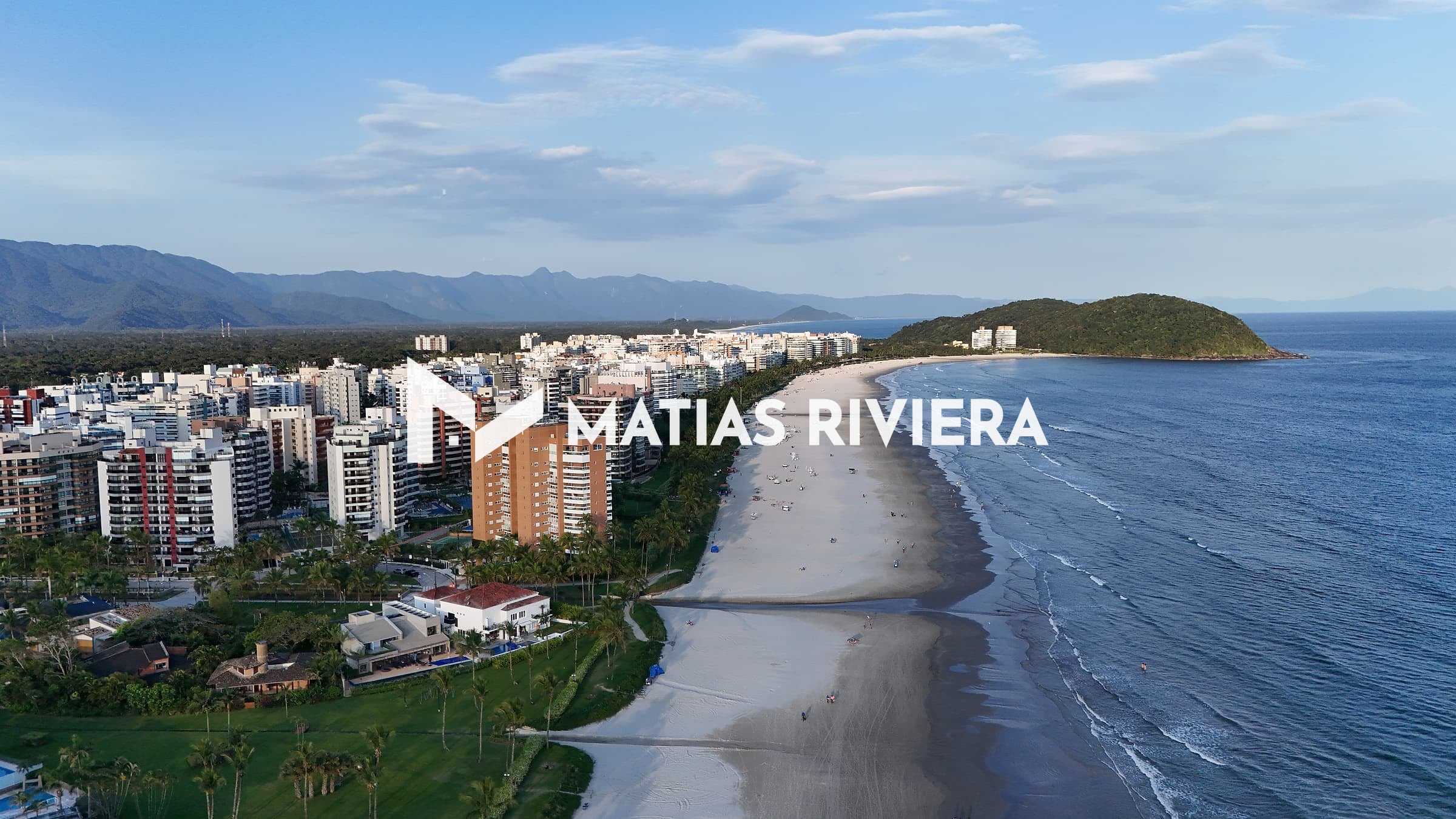 Vista aérea da orla da Riviera de São Lourenço no fim de tarde, com prédios e casas à esquerda, a faixa de areia em curva, o mar à direita e a serra ao fundo