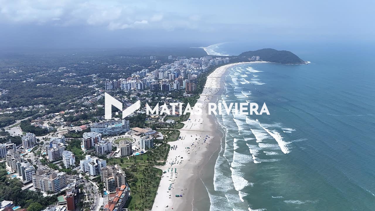 Vista aérea da orla da Riviera de São Lourenço com prédios e casas entre a vegetação, a faixa de areia com guarda-sóis, o mar com ondas e um morro no fim da praia