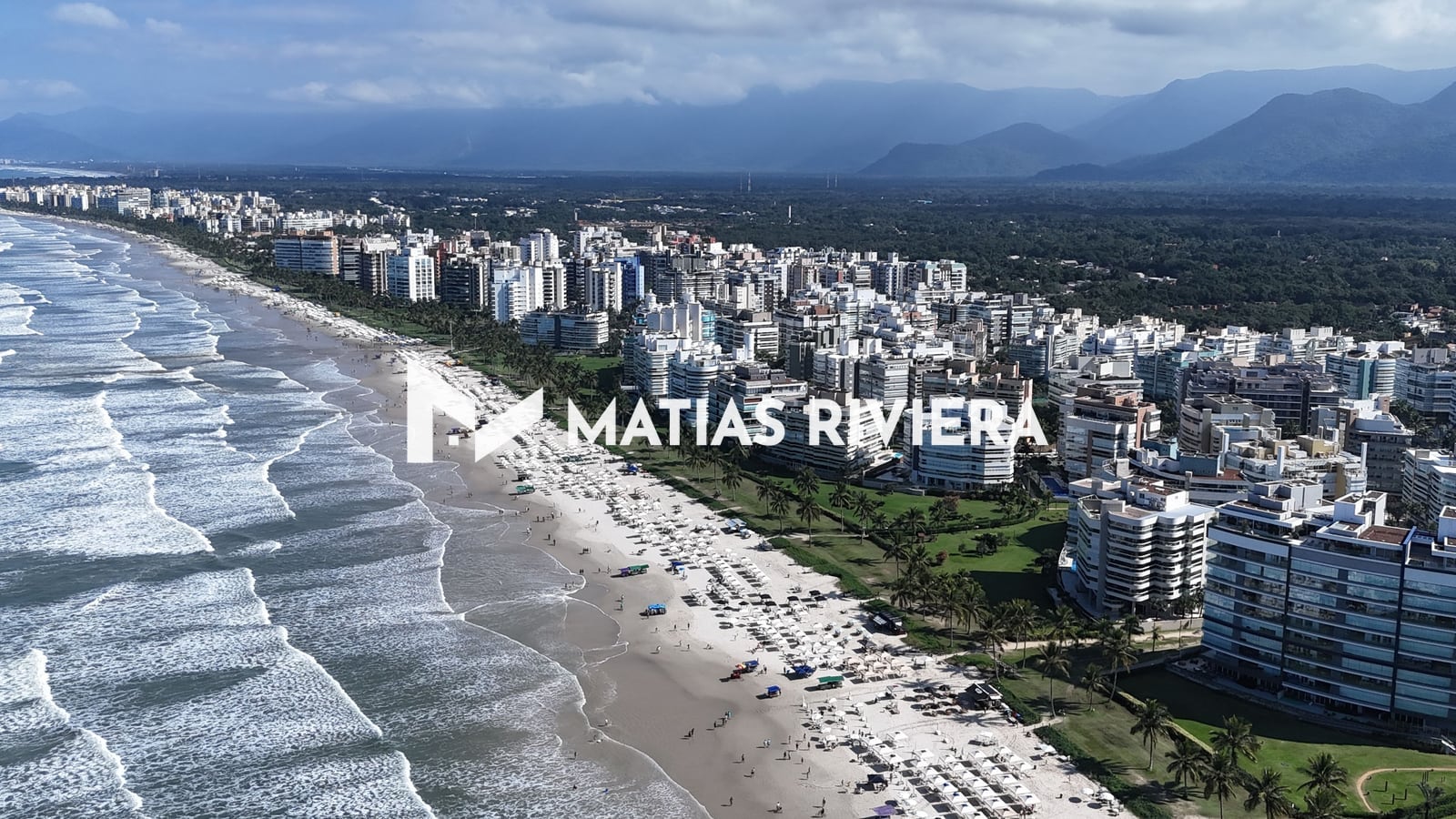 Faixa de areia da Riviera de São Lourenço com guarda-sóis, ondas e a serra ao fundo, em dia de sol