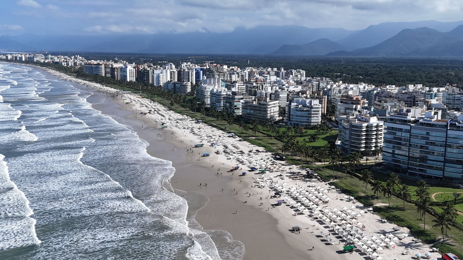Praia da Riviera de São Lourenço em dia de verão, com guarda-sóis na areia, ondas e os edifícios da orla