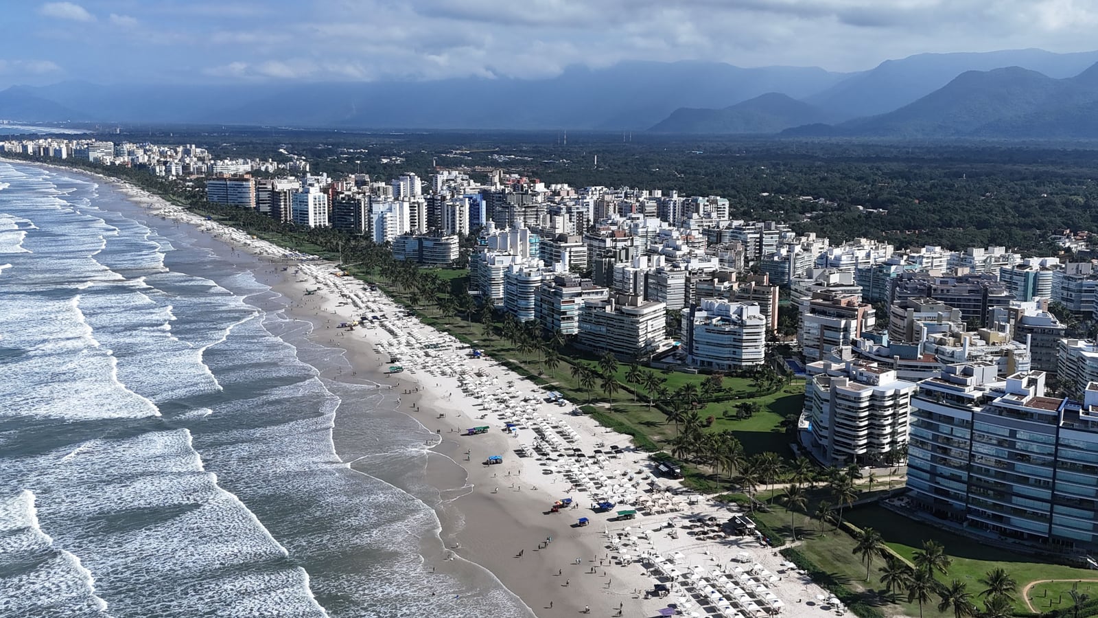 Faixa de areia da Riviera de São Lourenço com guarda-sóis, ondas e a serra ao fundo, em dia de sol