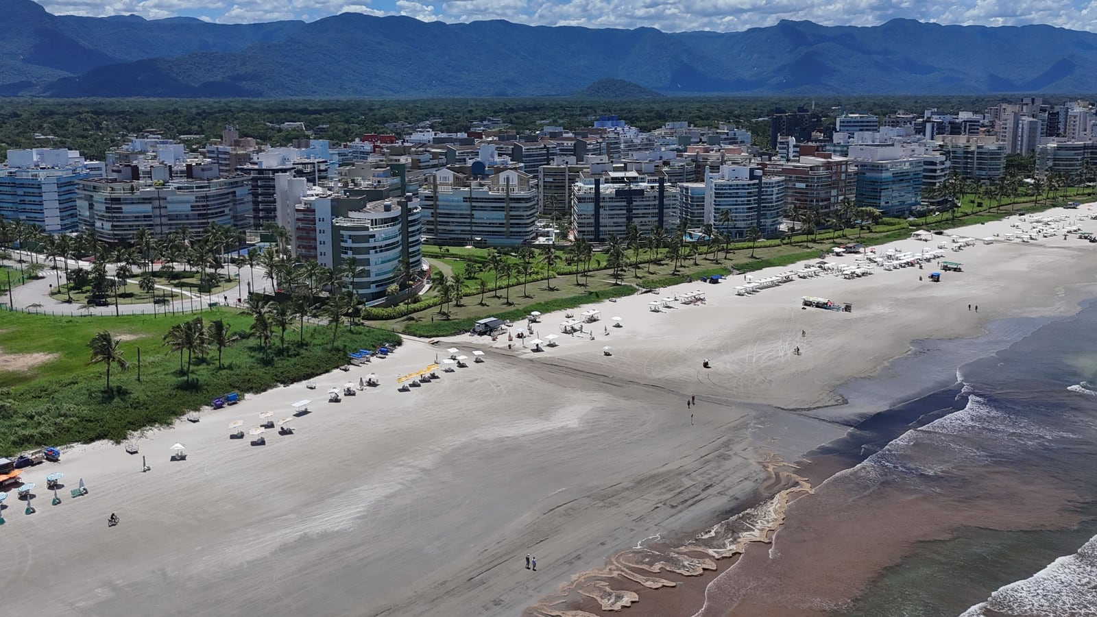 Faixa de areia larga da Riviera de São Lourenço com os edifícios da orla e a serra ao fundo, em dia de sol