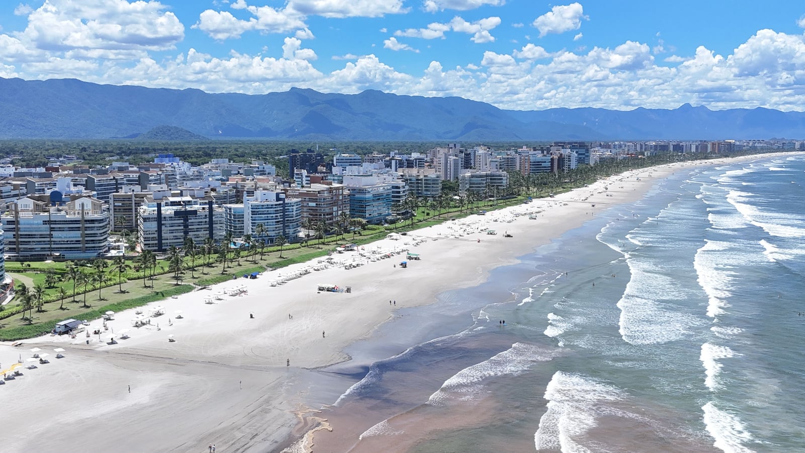 Praia da Riviera de São Lourenço em dia de sol, com a areia, o mar, os edifícios da orla e a serra ao fundo