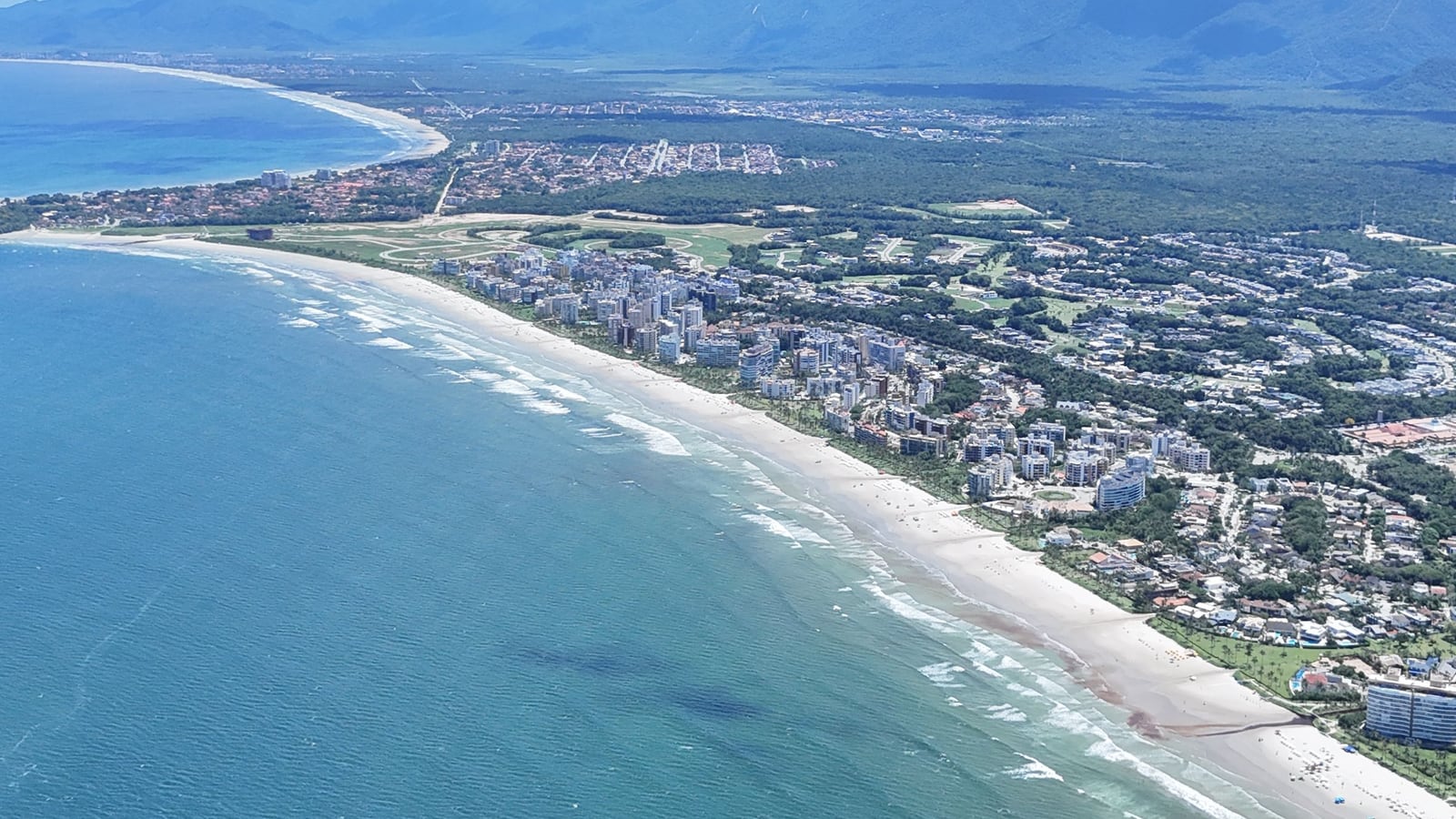 Mar azul e praia em curva da Riviera de São Lourenço, com os módulos residenciais e a serra no horizonte