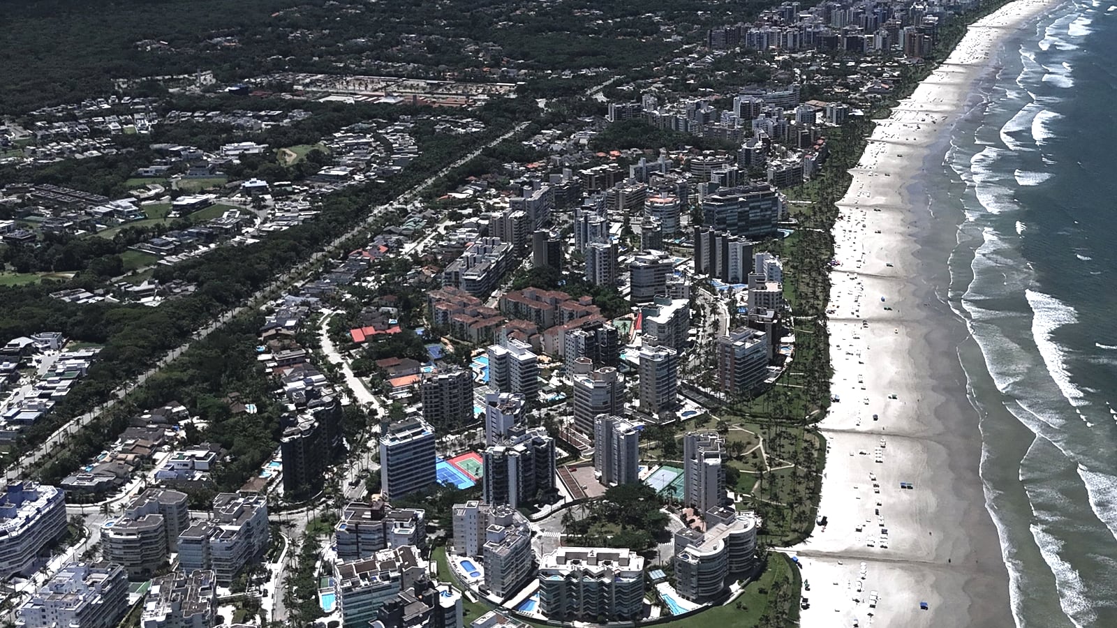 Edifícios e praia da Riviera de São Lourenço vistos do alto, com o morro e a Serra do Mar no horizonte