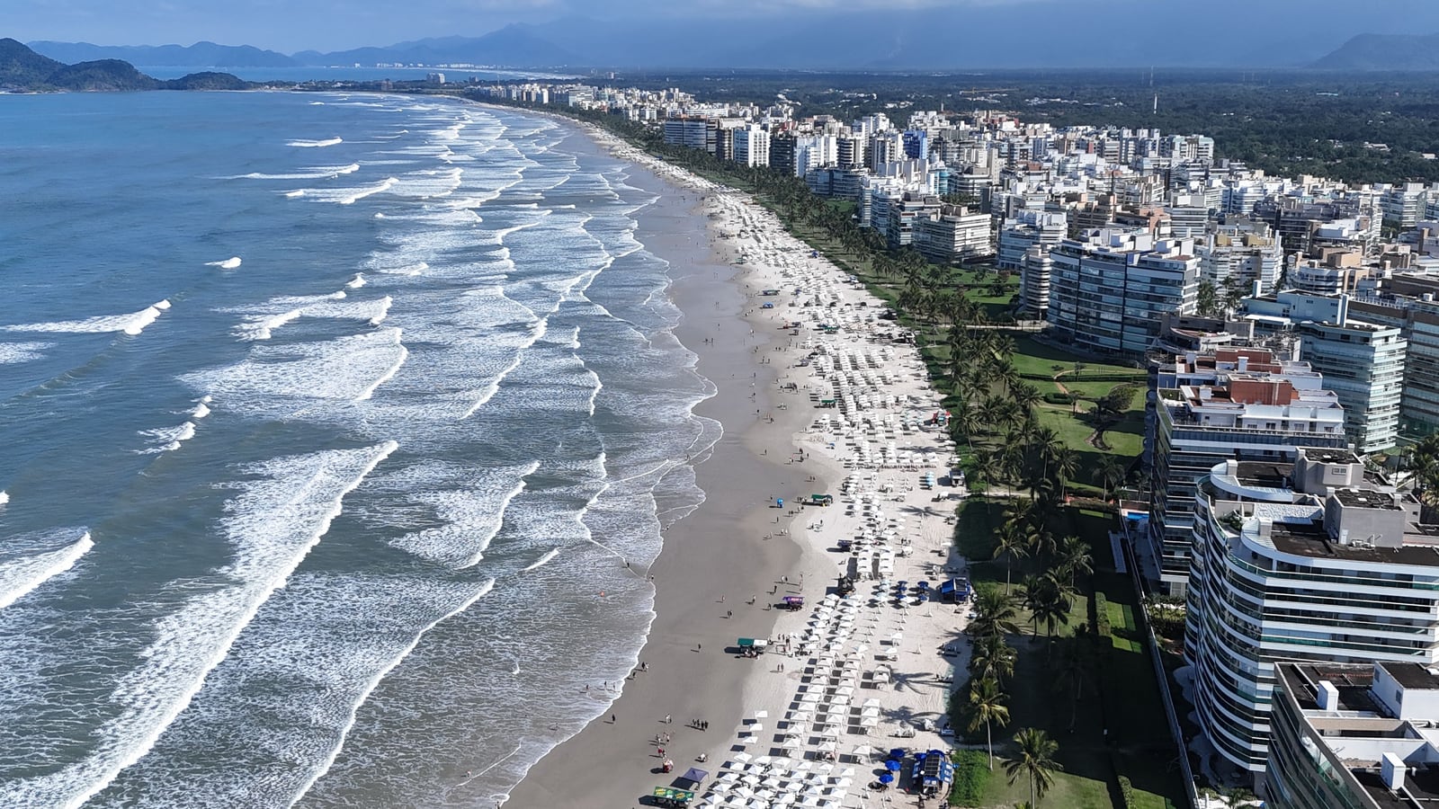 Orla da Riviera de São Lourenço com a ilha e a serra ao fundo, guarda-sóis na areia e ondas