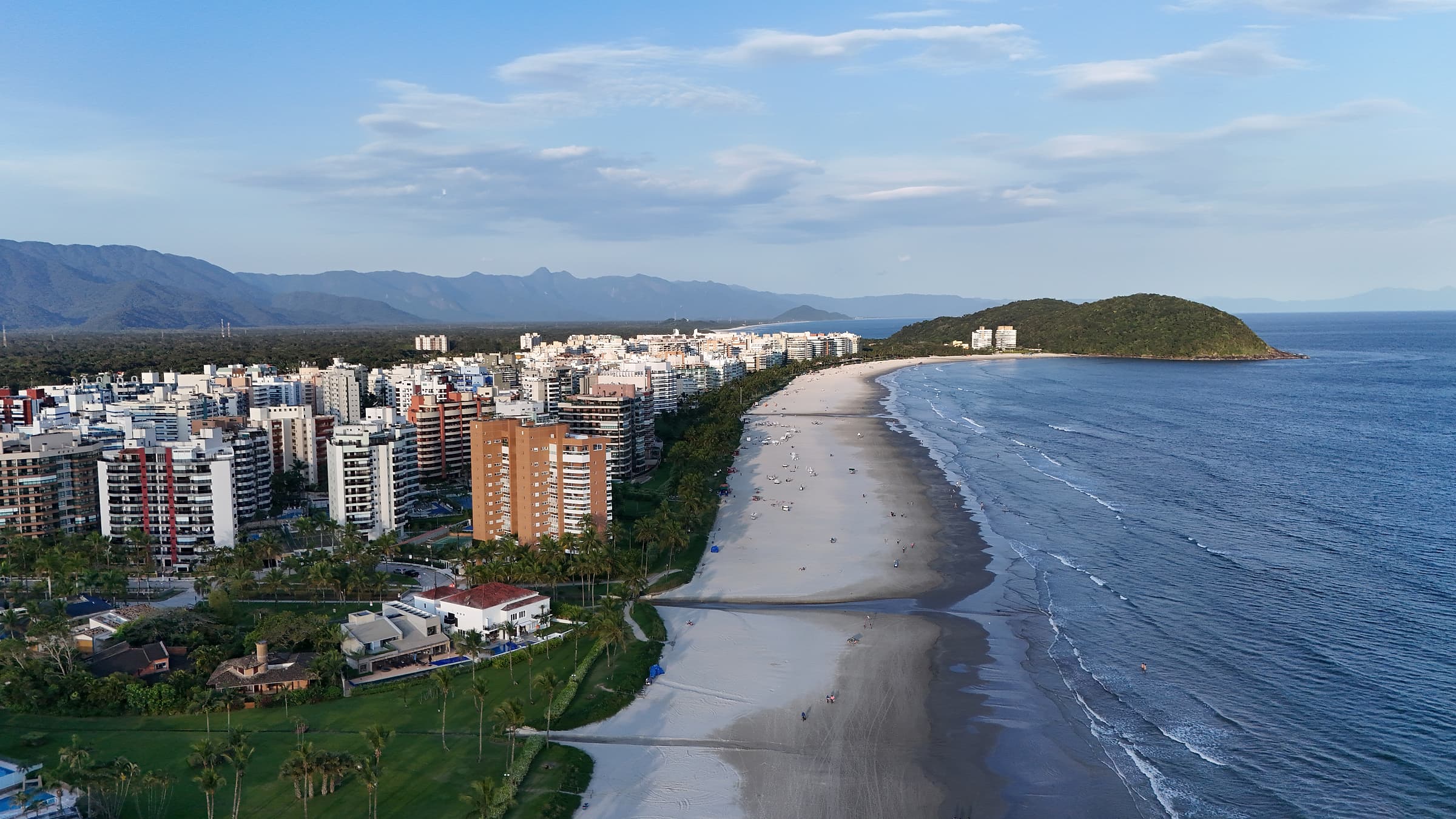 Vista aérea da orla da Riviera de São Lourenço no fim de tarde, com prédios e casas à esquerda, a faixa de areia em curva, o mar à direita e a serra ao fundo